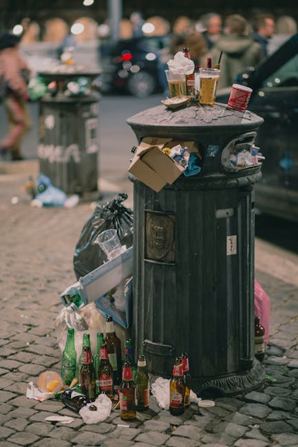 A public outdoor rubbish bin made of dark metal with a rounded lid, situated on a cobbled pavement. The bin is overflowing with various waste items, including clear plastic bottles, crumpled paper, and food containers, some of which have spilled onto the ground around it. On the top of the bin, there are disposable coffee cups with plastic lids, a paper food tray containing an ice cream cone, and a small cardboard box. Nearby, there are additional discarded bottles, crushed cans, and miscellaneous litter scattered around the base of the bin. In the background, blurred figures of people and parked cars suggest a busy urban setting. The scene exemplifies improper disposal and highlights the need for private waste management services such as those offered by Rubbish Removal Ruislip, which specialise in alternative waste handling solutions for local communities.