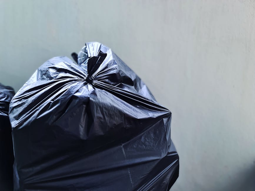 Close-up image of a tightly knotted black plastic rubbish bag with a shiny and slightly crinkled surface, resting against a plain, light-colored wall. The bag appears to be filled with waste material, causing it to bulge and form a rounded shape. The texture of the plastic reflects some light, highlighting the folds and creases. In the background, portions of additional black rubbish bags are visible, stacked or placed closely together, indicative of waste collection or disposal. The setting appears to be indoors or in a shaded outdoor area, with no other objects or environmental details visible. The scene conveys a typical scene associated with waste management, such as preparing refuse for collection by a rubbish removal service like Rubbish Removal Ruislip, supporting the context of private waste handling solutions.
